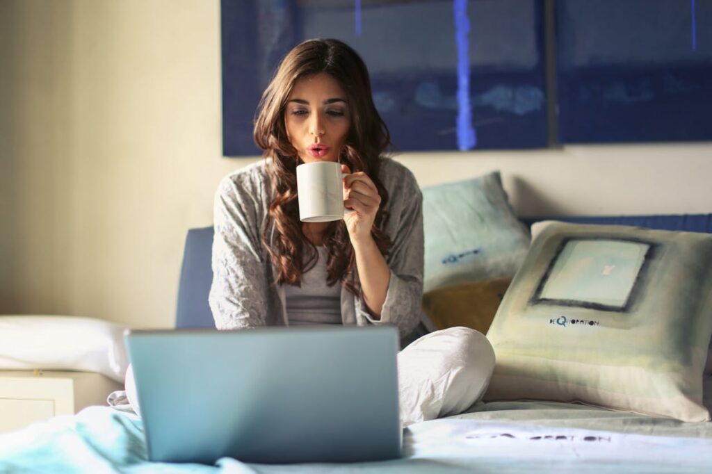 A woman sitting on her bed with legs crossed drinking a coffee.
