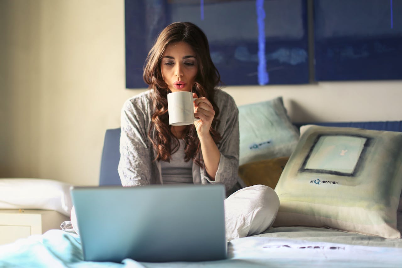 A woman sitting on her bed with legs crossed drinking a coffee.